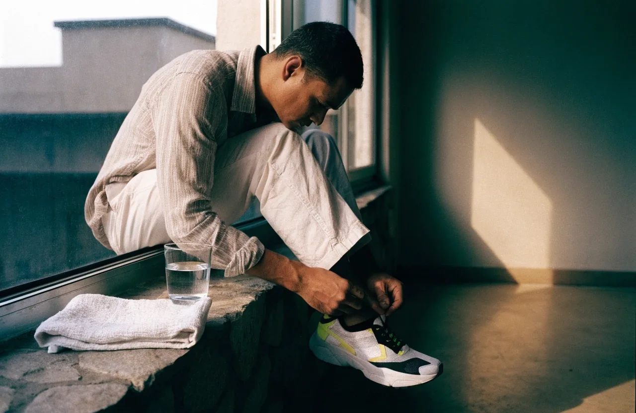 A man lacing a training shoe at a sunlit stone sill with a glass of water and a folded linen towel beside him